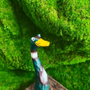 May include: A hand-carved wooden figurine of a mallard duck with a long, slender neck. The duck features a bright yellow beak, a dark green head, and a body painted with shades of brown, white, and green, showcasing visible wood grain texture.