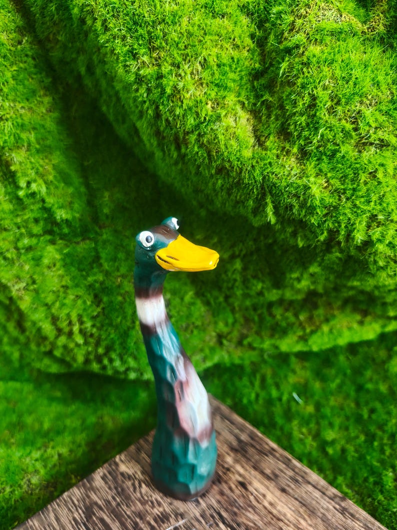 May include: A hand-carved wooden figurine of a mallard duck with a long, slender neck. The duck features a bright yellow beak, a dark green head, and a body painted with shades of brown, white, and green, showcasing visible wood grain texture.