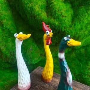 May include: Three hand-carved wooden figurines featuring a white duck, a yellow rooster with a red comb, and a green mallard duck. Each figure has a long, slender neck, a textured surface, and wide, cartoonish eyes, standing upright on a wooden base.