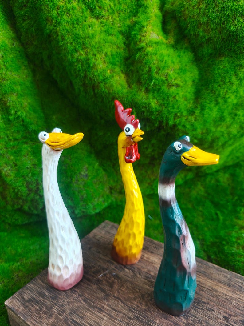May include: Three hand-carved wooden figurines featuring a white duck, a yellow rooster with a red comb, and a green mallard duck. Each figure has a long, slender neck, a textured surface, and wide, cartoonish eyes, standing upright on a wooden base.