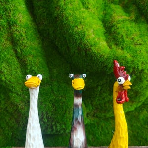 May include: Three hand-carved wooden figurines featuring elongated necks and wide, googly eyes. The left figure is white with a yellow beak, the center figure is dark green and brown with a yellow beak, and the right figure is yellow with a red comb.