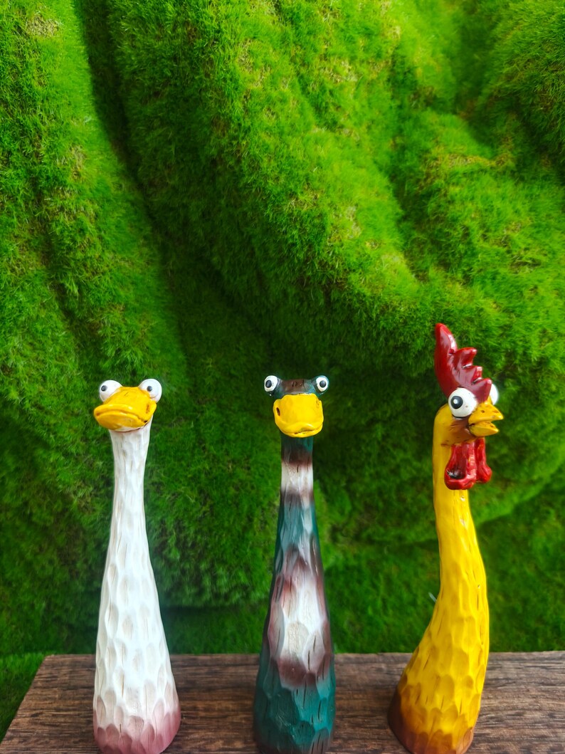 May include: Three hand-carved wooden figurines featuring elongated necks and wide, googly eyes. The left figure is white with a yellow beak, the center figure is dark green and brown with a yellow beak, and the right figure is yellow with a red comb.