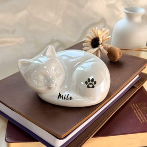 May include: A white ceramic cat ornament in a sleeping position, featuring the name "Milo" and a paw print. It sits on a stack of brown leather-bound books, with a small white vase and dried flower in the background.