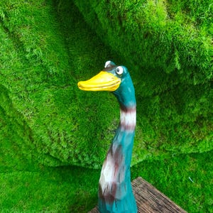 May include: A hand-carved wooden duck figurine featuring a long, slender neck painted in shades of teal, brown, and white. The duck has a bright yellow beak and two prominent, wide-set eyes, with a textured surface showing visible wood grain.