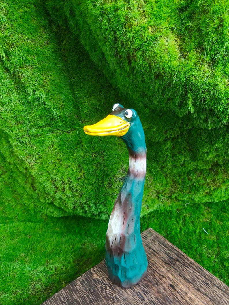 May include: A hand-carved wooden duck figurine featuring a long, slender neck painted in shades of teal, brown, and white. The duck has a bright yellow beak and two prominent, wide-set eyes, with a textured surface showing visible wood grain.