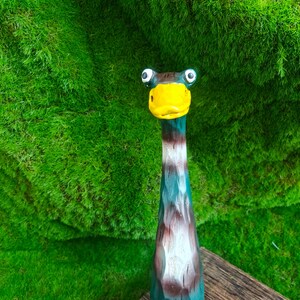 May include: A hand-carved wooden duck figurine with a long neck, featuring a bright yellow beak, white and dark brown painted details, and two prominent round eyes. The surface of the wood shows visible carved textures throughout the body.