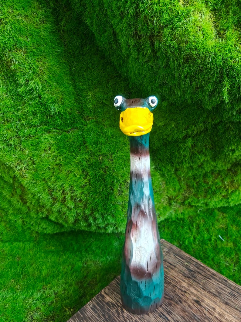 May include: A hand-carved wooden duck figurine with a long neck, featuring a bright yellow beak, white and dark brown painted details, and two prominent round eyes. The surface of the wood shows visible carved textures throughout the body.
