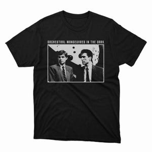 Może przedstawiać: Czarny t-shirt z czarno-białym zdjęciem dwóch mężczyzn w garniturach i napisem "ORCHESTRAL MANOEUVRES IN THE DARK" w białym prostokącie. T-shirt ma okrągły dekolt.