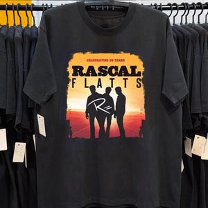 Può includere: T-shirt nero con la scritta "RASCAL FLATTS" e una silhouette di tre figure sullo sfondo di un tramonto. La maglietta presenta anche la scritta "CELEBRATING 25 YEARS". Il disegno è stampato su una maglietta nera a maniche corte.