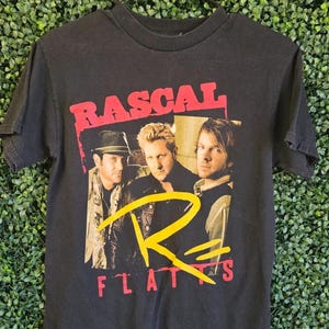 May include: Black t-shirt featuring a graphic of the band Rascal Flatts. The band's name is printed in red, with a yellow "R" and the word "FLATTS" in red. The image shows three band members.