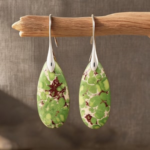 Peut inclure: Une paire de boucles d'oreilles en forme de goutte d'eau avec un motif tacheté vert et marron. Les boucles d'oreilles ont des crochets argentés et sont présentées sur une branche en bois. Un accessoire unique.