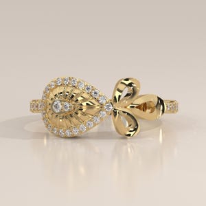 Può includere: Un anello in oro con un elemento centrale a forma di pera con un motivo a raggiera e due piccole pietre incastonate, circondato da un alone di piccole pietre bianche rotonde. La fascia è decorata con una fila di piccole pietre bianche e un motivo floreale.