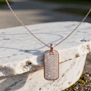 May include: Rose gold dog tag pendant necklace with a delicate chain. The rectangular pendant is encrusted with small, sparkling crystals. The necklace is displayed on a white stone surface.