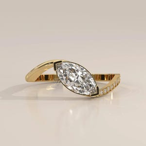 May include: A gold ring featuring a central marquise-cut clear gemstone held in a tension setting. The band is asymmetrical, with one side smooth and polished gold, while the other side is pavé-set with small round clear stones along the shank.