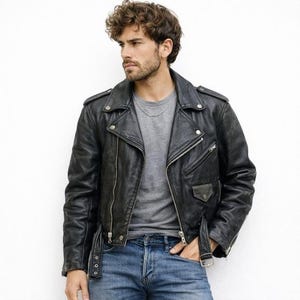 Puede incluir: Chaqueta de cuero negra de estilo motero con cremalleras y broches plateados. La chaqueta presenta un diseño asimétrico clásico, cuello con solapas y cinturón. El modelo lleva una camiseta gris y vaqueros azules.
