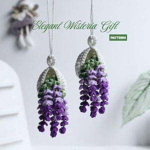 Op de afbeelding: Twee handgemaakte gehaakte wisteria bloemornamenten. De wisteria bloemen zijn paars en groen, met een witte gehaakte mand. De tekst "Elegant Wisteria Gift" is cursief. Het woord "PATTERN" staat in een groene doos.