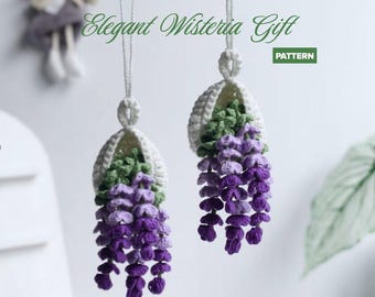 Może przedstawiać: Dwie wiszące ozdoby szydełkowe z białymi koszyczkami zawierającymi zielone liście i kaskadowe fioletowe kwiaty glicynii. Tekst Elegant Wisteria Gift PATTERN jest wyświetlany na zielono. Każda ozdoba jest zawieszona na białej pętli ze sznurka.