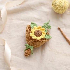 Puede incluir: Un ramo de ganchillo hecho a mano con un girasol amarillo con centro marr&oacute;n y hojas verdes. El ramo est&aacute; en un soporte marr&oacute;n en forma de cesta, atado con un lazo verde. Tambi&eacute;n se ve una madeja de lana amarilla y un ganchillo.