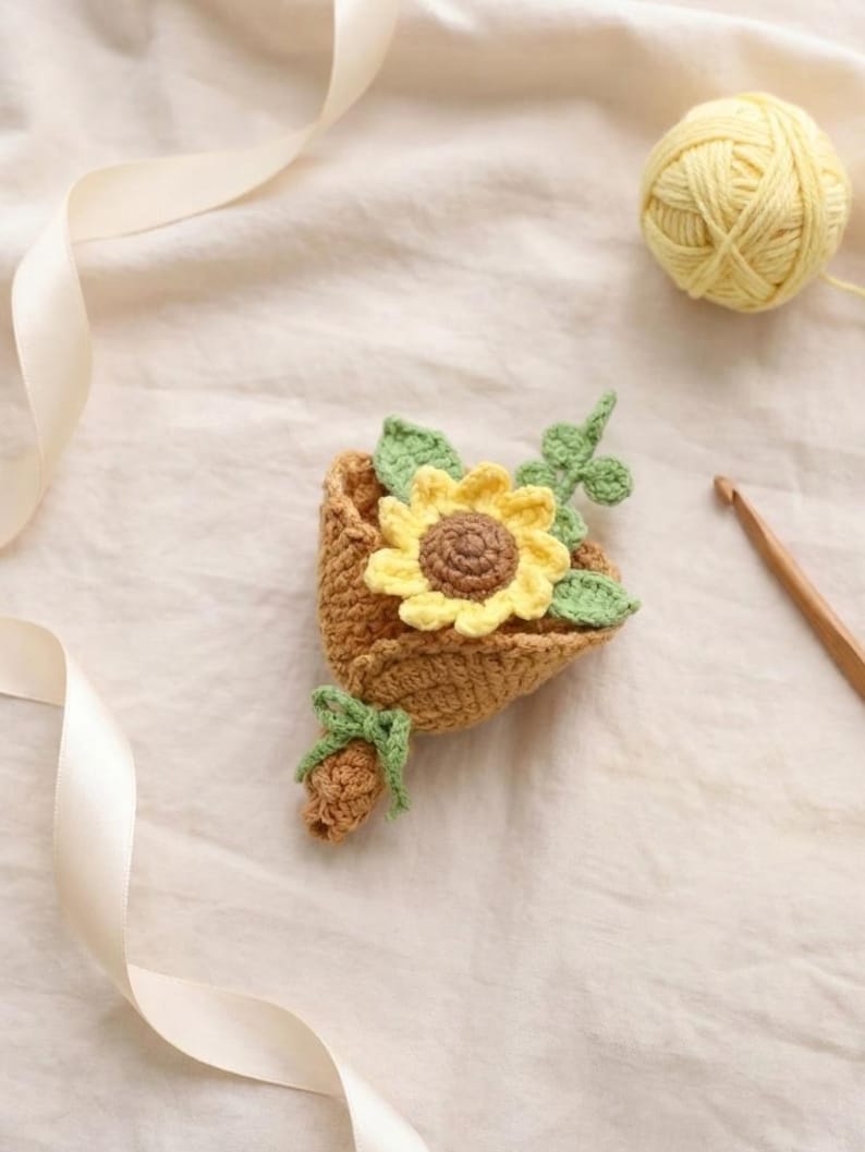 Puede incluir: Un ramo de ganchillo hecho a mano con un girasol amarillo con centro marr&oacute;n y hojas verdes. El ramo est&aacute; en un soporte marr&oacute;n en forma de cesta, atado con un lazo verde. Tambi&eacute;n se ve una madeja de lana amarilla y un ganchillo.