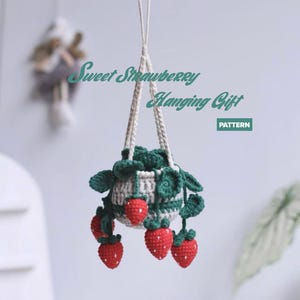 May include: A crocheted hanging plant featuring a white and green pot with red strawberries and green leaves, suspended by white yarn cords. The text Sweet Strawberry Hanging Gift PATTERN is visible in green and white against the light background.
