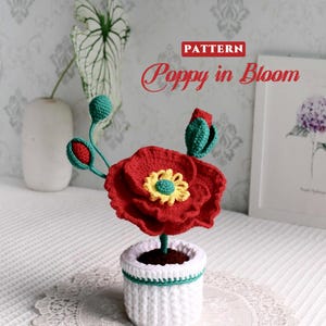 May include: A handmade crochet poppy flower in a white pot with a green trim. The red flower has a yellow center. The text "PATTERN Poppy in Bloom" is visible in the image. The flower is on a white lace doily.