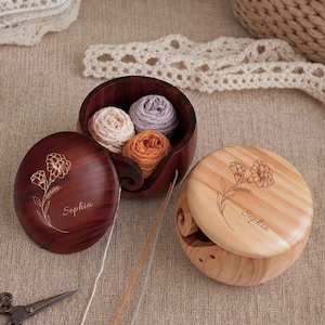May include: Two round wooden yarn bowls with lids, one dark brown and one light tan. Each lid features an engraved floral design and the name Sophia. The dark bowl contains three small balls of yarn in cream, lavender, and orange colors.