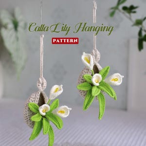 May include: Two crocheted calla lily hanging planters. Each planter features white calla lilies with yellow centres and green leaves, set in a beige crocheted basket. The text "Calla Lily Hanging PATTERN" is displayed on the image.