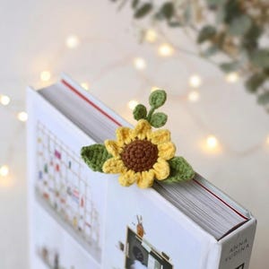 Puede incluir: Un marcap&aacute;ginas de girasol tejido a crochet con p&eacute;talos amarillos y centro marr&oacute;n, acentuado por hojas verdes. El marcap&aacute;ginas est&aacute; colocado en la esquina de un libro. El libro tiene una cubierta blanca con el texto "FURNITURE" y el nombre del autor.