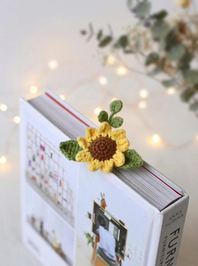 Puede incluir: Un marcap&aacute;ginas de girasol tejido a crochet con p&eacute;talos amarillos y centro marr&oacute;n, acentuado por hojas verdes. El marcap&aacute;ginas est&aacute; colocado en la esquina de un libro. El libro tiene una cubierta blanca con el texto "FURNITURE" y el nombre del autor.