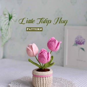 May include: A crocheted tulip arrangement in a small pot. The arrangement features three pink tulips with green stems and leaves. The pot is beige with a pink trim, and the text "Little Tulip Hug PATTERN" is visible.