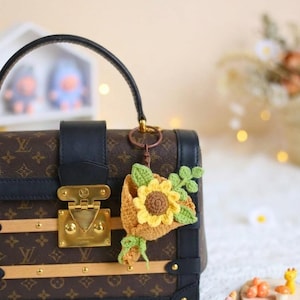 Puede incluir: Un bolso marr&oacute;n y negro con cierre y asa dorados, adornado con un llavero de girasol de ganchillo. El llavero presenta un girasol amarillo con hojas verdes y una cesta marr&oacute;n.