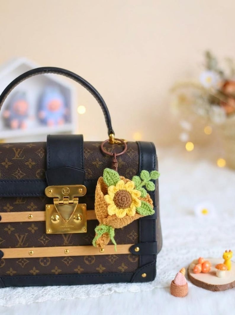 Puede incluir: Un bolso marr&oacute;n y negro con cierre y asa dorados, adornado con un llavero de girasol de ganchillo. El llavero presenta un girasol amarillo con hojas verdes y una cesta marr&oacute;n.