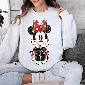 May include: A light gray sweatshirt featuring a graphic of Minnie Mouse. Minnie is depicted in a seated pose, with a red bow with white polka dots. The graphic is in black, red, and white.
