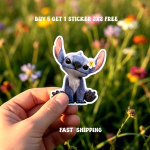 May include: A blue cartoon character sticker with a white flower on its ear. The sticker is held in a hand against a blurred background of flowers. Text on the sticker reads "BUY 5 GET 1 STICKER 2X2 FREE".