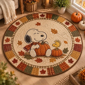 May include: A round quilted rug featuring a cream-colored center with an illustration of Snoopy wearing a scarf and holding a pumpkin, alongside Woodstock sitting on a smaller pumpkin, surrounded by a border of autumn leaves and plaid squares.