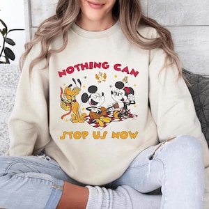 May include: Cream-colored sweatshirt featuring a graphic of Mickey Mouse, Minnie Mouse, and Pluto on a picnic blanket. The text "NOTHING CAN STOP US NOW" is printed in red and yellow. The design includes stars and bees.