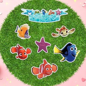 May include: A collection of colorful cartoon fish stickers, including clownfish, a blue tang, and a sea turtle, arranged on a green, circular surface. A banner with more sea creatures is at the top. The background is a pink surface.