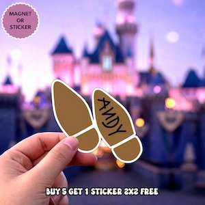 May include: Two brown boot-shaped stickers or magnets with a white border. One boot is plain brown, and the other features the name ANDY written in black capital letters. The items are held against a blurred background of a castle at twilight.