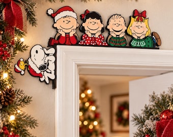 Może przedstawiać: Dekoracja narożna drzwi z motywem Peanuts przedstawiająca Charliego Browna, Lucy, Linusa i Sally opierających brody na dłoniach wzdłuż górnej ramy oraz Snoopy'ego i Woodstocka w czapkach Mikołaja wiszących na bocznej ramie.