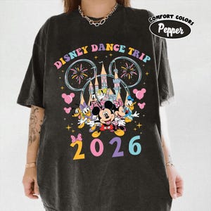 May include: Dark gray t-shirt featuring a colorful graphic of Disney characters, fireworks, and a castle. The text "DISNEY DANCE TRIP" arches across the top, with "2026" in large, colorful numbers at the bottom. The shirt has a Comfort Colors Pepper label.