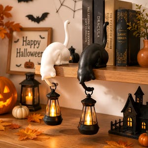 May include: Two cat-shaped shelf sitters, one white and one black, each featuring a long tail that curves downward to hold a small black lantern with a glowing orange light. The cats are positioned on the edge of a wooden shelf.