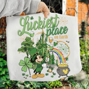 May include: White tote bag with a St. Patrick's Day theme. It features Mickey Mouse in green attire, a castle, rainbow, and a pot of gold. The text says "Luckiest place on Earth."