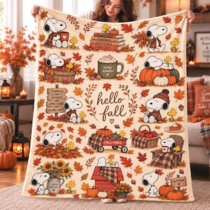 May include: A cream-colored throw blanket featuring a repeating pattern of Snoopy and Woodstock in autumn scenes. Illustrations include pumpkins, falling leaves, sunflowers, a red wagon, and mugs with text like 'cozy season' and 'hello fall'.