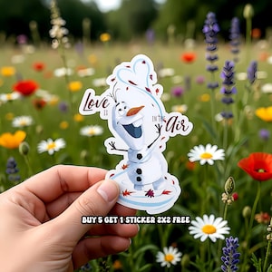 Puede incluir: Una pegatina con Olaf de Frozen, con el texto "Love is in the Air". Olaf es blanco con una nariz de zanahoria y una gran sonrisa. La pegatina se sostiene frente a un campo de flores coloridas.