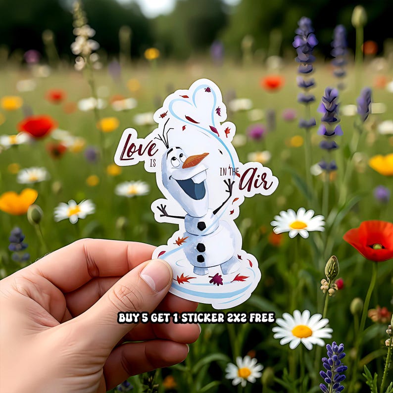 Puede incluir: Una pegatina con Olaf de Frozen, con el texto "Love is in the Air". Olaf es blanco con una nariz de zanahoria y una gran sonrisa. La pegatina se sostiene frente a un campo de flores coloridas.