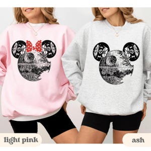 May include: Two sweatshirts, one light pink and one ash, each featuring a graphic of a Death Star with Mickey Mouse ears. The light pink sweatshirt has a red polka dot bow. The ash sweatshirt has a similar design without the bow. The words "light pink" and "ash" are at the bottom.