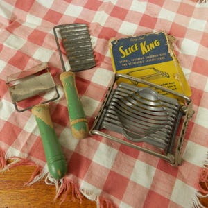 May include: A collection of vintage kitchen tools, including a metal egg slicer, graters, and a corrugated cutter with green wooden handles. The tools are arranged on a red and white checkered cloth, with the original "Slice King" packaging.
