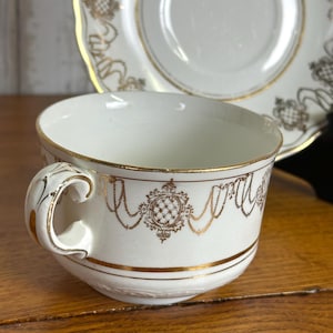 May include: White teacup and saucer set with gold filigree detailing. The teacup has a rounded shape with a handle and gold trim. The saucer is square with rounded edges and gold accents. The set is likely vintage or antique.