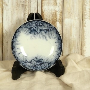 May include: A small, round, antique plate with a white center and a blue floral design around the rim. The plate is displayed on a black stand against a light wood background. The floral pattern includes leaves and flowers.