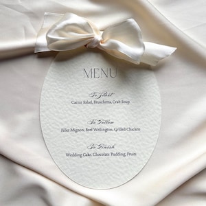 Puede incluir: Un menú ovalado con un lazo de seda color crema. El menú, con la palabra «MENU» en la parte superior, enumera entrantes, platos principales y postres, incluyendo ensalada César, filete mignon y tarta de boda.